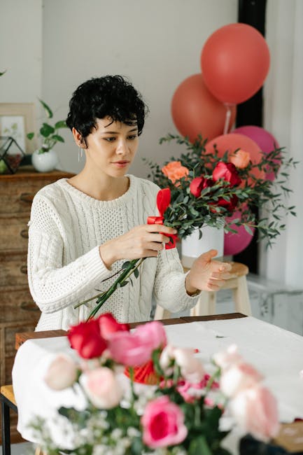 A young woman with short, curly black hair and light skin is carefully arranging a floral gift consisting of bright pink and red roses, accented with greenery, tied together with a red ribbon. She is 