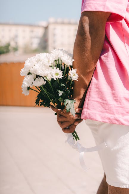 A person in a pink top and white shorts holds a bouquet of fresh white chrysanthemums and daisies, wrapped with a white ribbon, behind their back. The bouquet features vibrant green stems and leaves, 