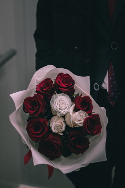 A person dressed in dark clothing, possibly a suit, holding a bouquet of fresh roses. The bouquet features a mix of deep red and cream roses arranged in a circular, floral design, wrapped in white pap