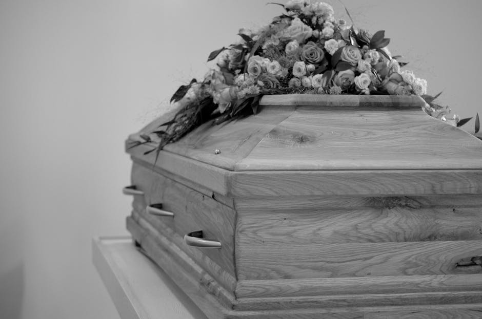 A wooden casket with a polished finish rests on a white table, adorned with a floral arrangement on top. The bouquet features a variety of flowers in soft shades, including roses, carnations, and fill