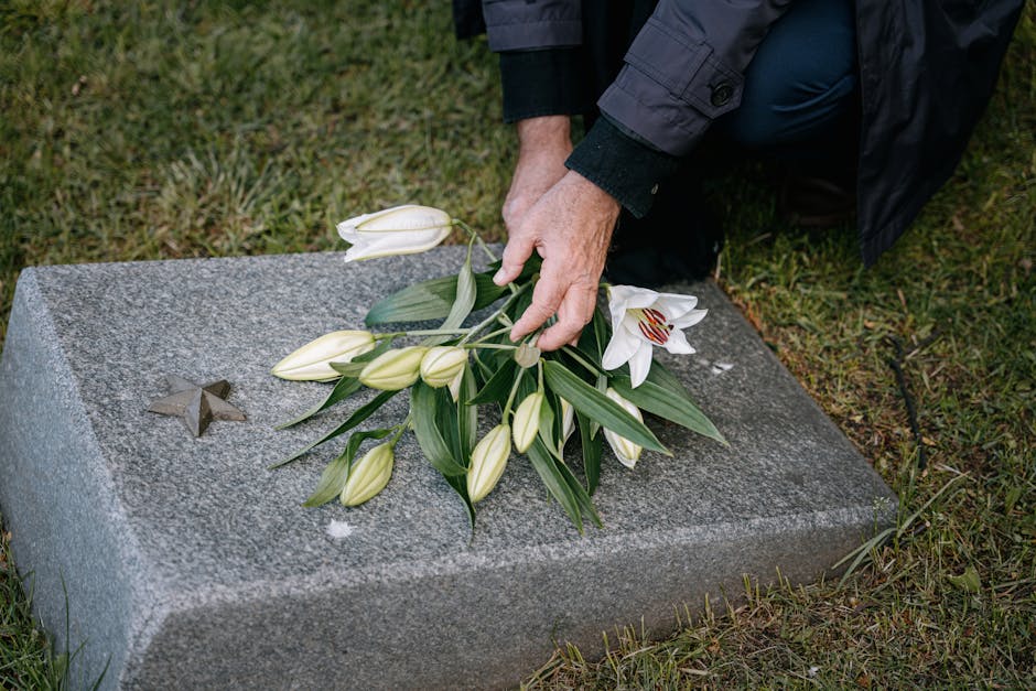 A person placing a floral arrangement of white lilies and unopened lily buds, with green leaves, on a gray granite memorial gravestone featuring a small star-shaped engraving. The arrangement is wrapp