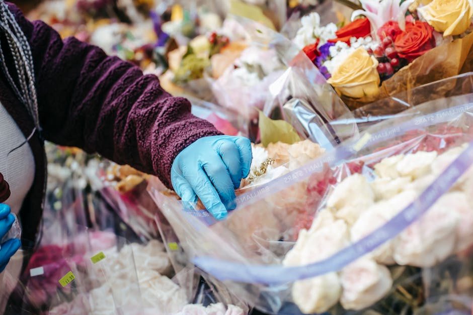 A close-up of a florist wearing a burgundy velvet long-sleeve top and blue disposable gloves, carefully handling a bouquet of fresh flowers wrapped in clear plastic. The arrangement includes cream, pe