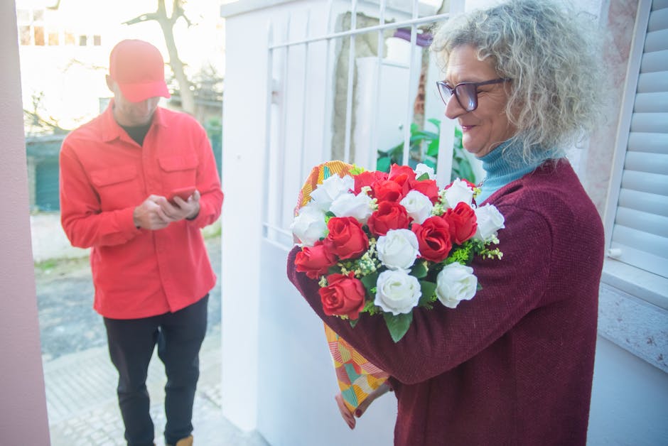 A woman with curly gray hair and glasses, wearing a burgundy sweater over a blue turtleneck, is holding a vibrant bouquet of fresh red and white roses wrapped in multicolored paper. She stands near a 