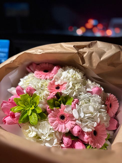 A bouquet of fresh pink gerbera daisies, white hydrangeas, and green foliage arranged together and wrapped in brown kraft paper, likely prepared for flower delivery. The flowers display vibrant pink p