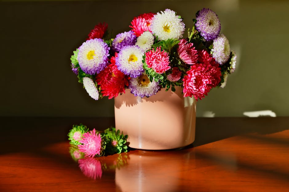 A vibrant floral bouquet featuring a mix of purple, pink, and white chrysanthemums arranged in a soft pink ceramic vase. The flowers display full, fresh petals with a textured appearance, and some ste