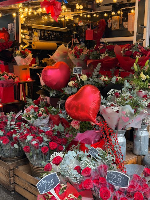 A vibrant display of fresh flowers featuring red roses, white baby's breath, and green foliage arranged in bouquets and wraps. The flowers include long-stemmed roses in clear plastic wrapping, and sma