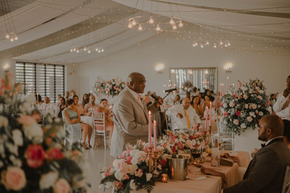A floral arrangement featuring pink and white roses, peonies, and assorted greenery, placed on a table with tall pink candles in gold holders. The arrangement is lush and full, with flowers in various